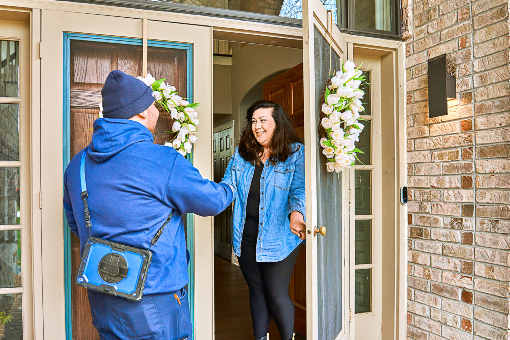 The woman opens the front door and shakes hands with the man in a blue uniform carrying a tool bag, who is prepared to service her heating system.