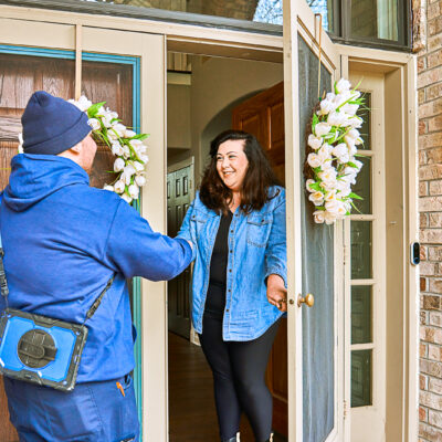 The woman opens the front door and shakes hands with the man in a blue uniform carrying a tool bag, who is prepared to service her heating system.