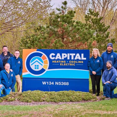 People in blue jackets pose around a Capital Heating, Cooling, and Electric sign in a grassy area with trees in the background, showcasing their expertise in maintaining year-round comfort.