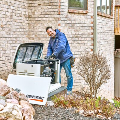 A man in a blue jacket expertly repairs a Generac generator beside a brick house, using Milwaukee tools to guarantee smooth and cool operation.