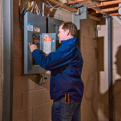 A person in a blue jacket meticulously examines an electric circuit breaker panel in a Milwaukee basement, surrounded by exposed pipes and wiring.