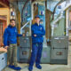 Wearing blue uniforms, two technicians stand in front of large HVAC systems in a basement. One technician points at the equipment while they discuss the complex electric components that control both heating and cooling functions.