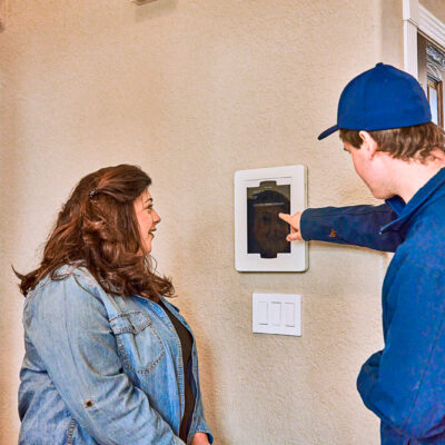 A person in a blue uniform demonstrates the functions of an electric wall-mounted device to a woman by showing her the screen.