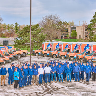 People in blue uniforms stand in a parking lot surrounded by orange and blue heating and cooling vans. Trees and a building frame this typical scene from Milwaukee.