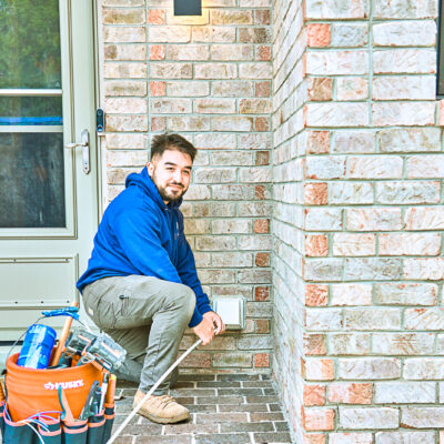 A person in a blue hoodie and work boots kneels by a brick wall and holds electric tools. A tool bag sits nearby on the brick pathway.