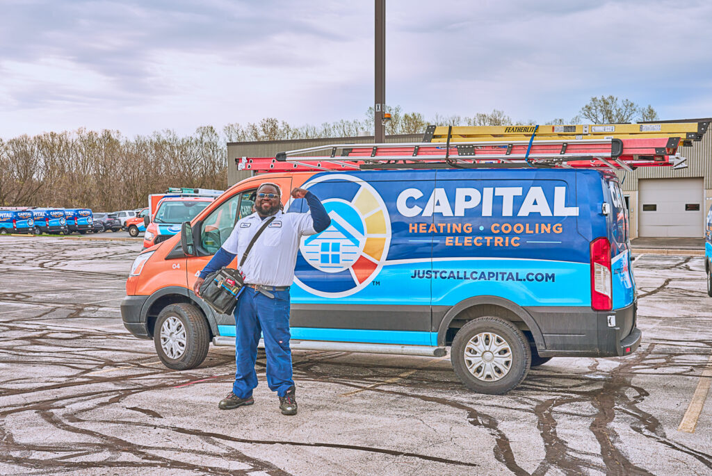 A worker gives a thumbs up and holds a tool bag while standing in front of a Capital Heating, Cooling, and Electric van in a Milwaukee parking lot.