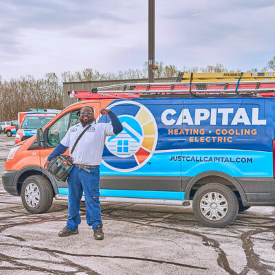 A worker gives a thumbs up and holds a tool bag while standing in front of a Capital Heating, Cooling, and Electric van in a Milwaukee parking lot.