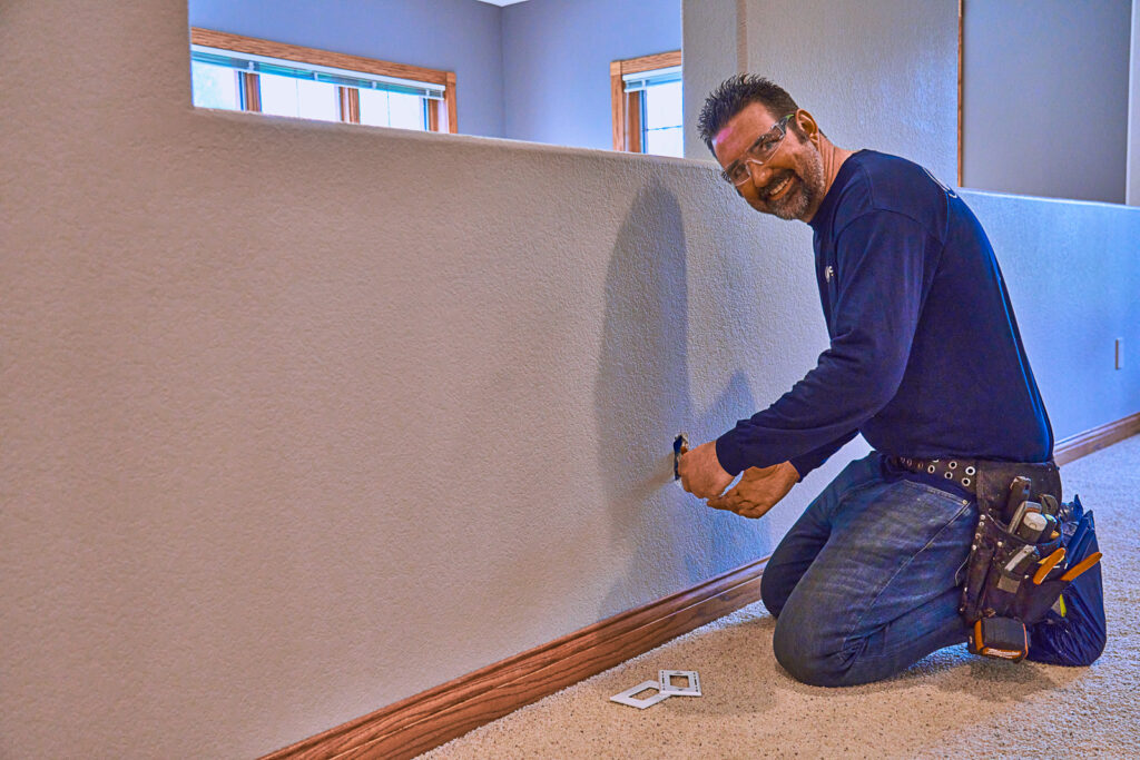 The man kneels on the floor and skillfully works on an electric wall outlet with a screwdriver. Wearing jeans, a long-sleeve shirt, and a tool belt, he manages the task in the well-lit room with two windows, ensuring everything is ready for Milwaukee's chilly days.