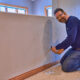 The man kneels on the floor and skillfully works on an electric wall outlet with a screwdriver. Wearing jeans, a long-sleeve shirt, and a tool belt, he manages the task in the well-lit room with two windows, ensuring everything is ready for Milwaukee's chilly days.