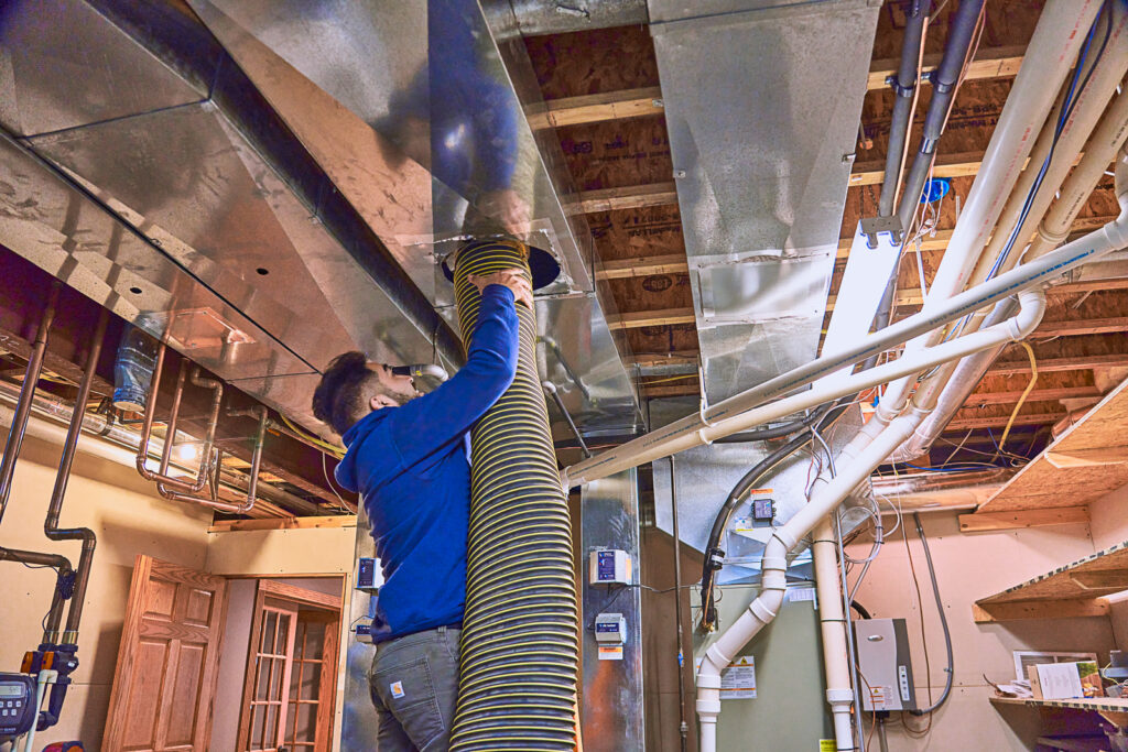 A professional diligently cleans the ducts in a basement, using a large hose connected to the ductwork.