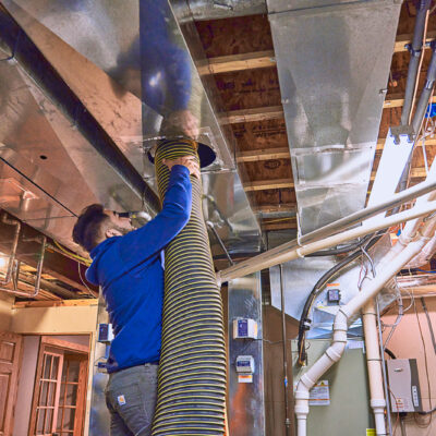 A professional diligently cleans the ducts in a basement, using a large hose connected to the ductwork.