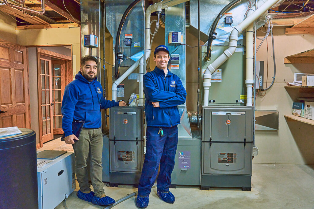 Two technicians in blue uniforms inspect a potential furnace water leak next to large HVAC units in a basement.