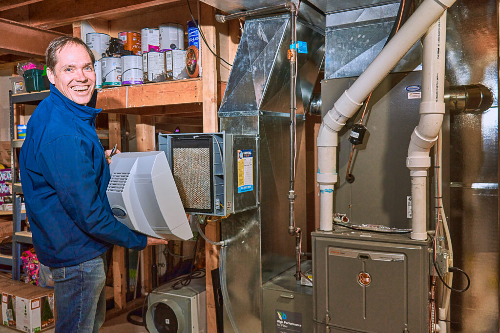 A person installs a heating filter in a basement furnace, while shelves filled with paint cans stand in the background.