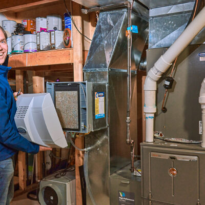 A person installs a heating filter in a basement furnace, while shelves filled with paint cans stand in the background.