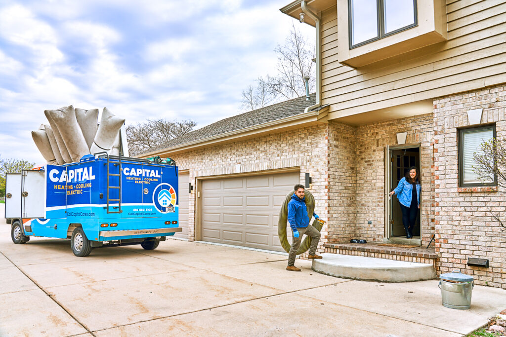 A technician carries large pipes from a bright blue Capital Heating & Cooling truck to a brick house.