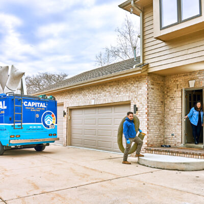 A technician carries large pipes from a bright blue Capital Heating & Cooling truck to a brick house.