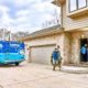 A technician carries large pipes from a bright blue Capital Heating & Cooling truck to a brick house.