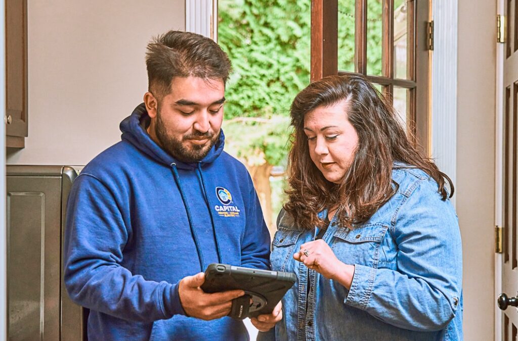 Indoors, two people intensely focus on a tablet titled "Everything You Need to Know." One person in a blue hoodie with a logo and another in a denim jacket explore this Boilers Guide. A doorway and greenery in the background enhance the serene setting.