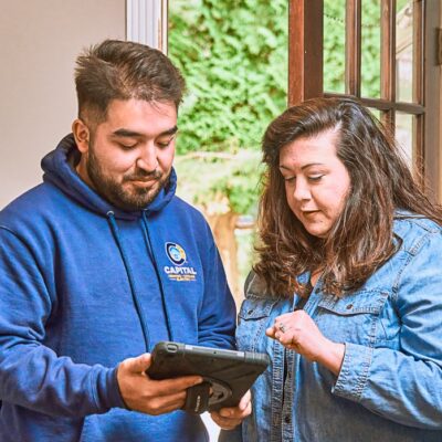 Indoors, two people intensely focus on a tablet titled "Everything You Need to Know." One person in a blue hoodie with a logo and another in a denim jacket explore this Boilers Guide. A doorway and greenery in the background enhance the serene setting.