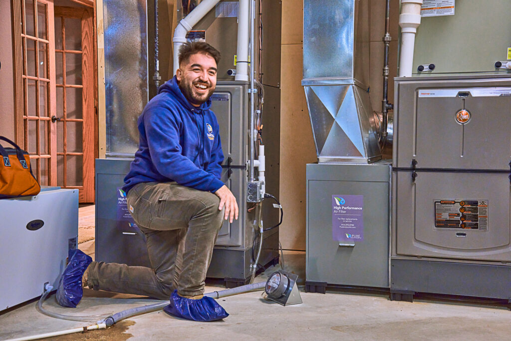 A person in a blue hoodie and shoe covers kneels in front of the furnace and beams at the camera, showcasing the art of Furnace Beyondification.