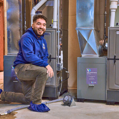 A person in a blue hoodie and shoe covers kneels in front of the furnace and beams at the camera, showcasing the art of Furnace Beyondification.