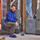 A person in a blue hoodie and shoe covers kneels in front of the furnace and beams at the camera, showcasing the art of Furnace Beyondification.