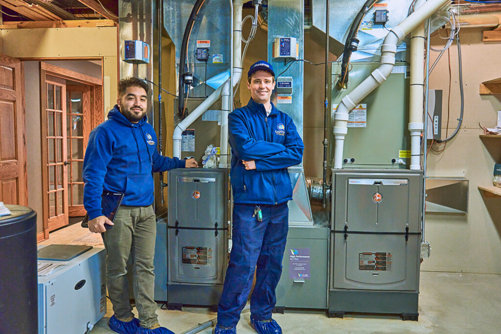 Two technicians in blue uniforms stand in front of heating and cooling systems, including a furnace, in a basement. They are ready to maintain the equipment and ensure everything runs smoothly for the upcoming winter season.